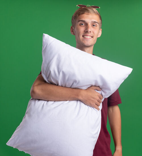 枕头微笑着的年轻帅哥穿着红衬衫 抱着被隔离在绿墙上的枕头穿着拥抱微笑