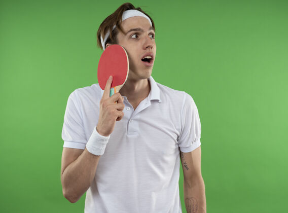 脸惊讶的年轻人一边看着戴着头带的腕带一边把乒乓球拍放在隔离在绿色墙上的脸上球拍运动壁板