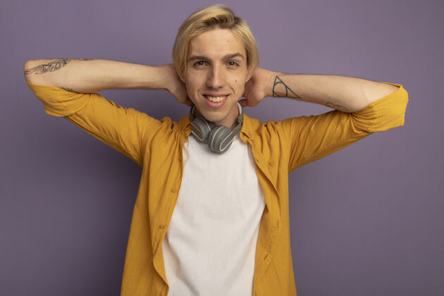 抱着微笑着的年轻金发男子身穿黄色t恤 脖子上戴着耳机 手放在脖子上 孤立在紫色上手紫色戴着