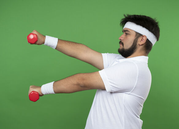 男站在侧视图年轻的运动男子戴着头带和腕带锻炼与哑铃隔离在绿色的墙壁佩戴腕带运动型