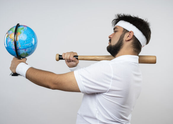 运动年轻的运动型男子戴着头带和腕带站在侧视图中 把棒球棒放在肩膀上 把地球仪放在隔离在白色墙上的一边男子年轻肩膀