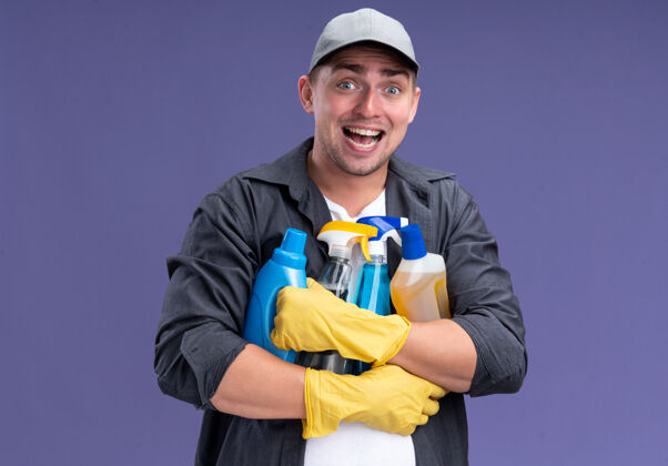 帽子兴奋的年轻帅哥 穿着t恤 戴着帽子和手套 手里拿着清洁工具 隔离在紫色的墙上小伙子兴奋手套