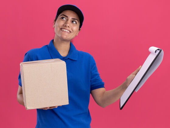 交货令人印象深刻的一面看年轻的送货女孩穿着制服与帽子举行与剪贴板在粉红色墙上孤立的盒子举行剪贴板年轻