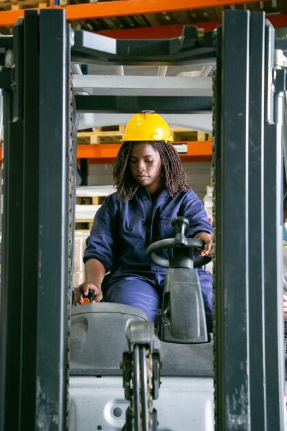 信心专注的年轻女物流员戴着安全帽在仓库里操作叉车 拉杠杆机器车轮女性