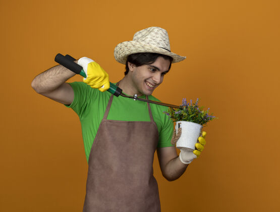 花年轻的男园丁面带微笑 穿着制服 戴着园艺帽 戴着手套 拿着剪子 在桔黄色的花盆里切花园艺橘子手套