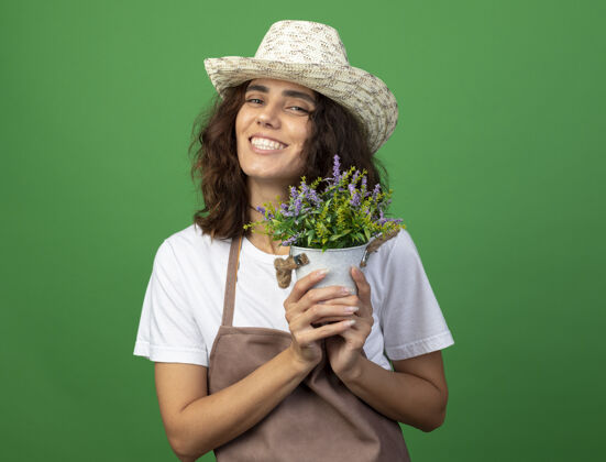 花园微笑着的年轻女园丁穿着制服戴着园艺帽捧着花盆里的花隔离在绿色上制服花花盆