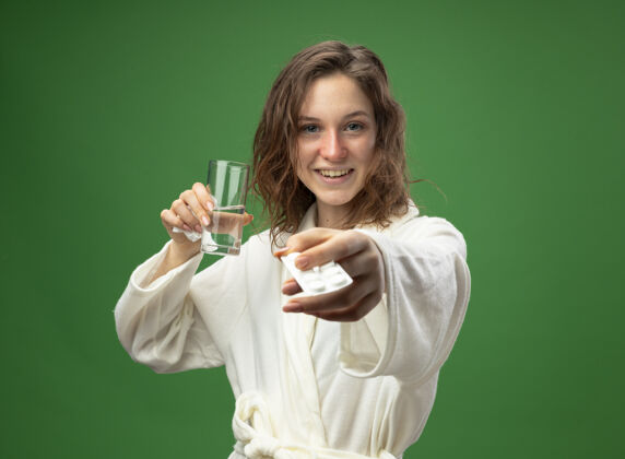 水微笑着的年轻病女孩穿着白色长袍 手里拿着一杯水 手里拿着隔离在绿色上的药丸药丸疾病绿色