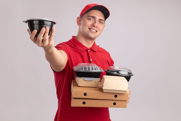 制服微笑着的年轻送货员穿着制服 戴着帽子 拿着比萨饼盒上的食品容器 拿着隔离在白墙上的食品容器食物盒子容器