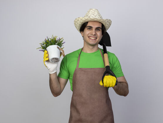 抱身着制服 面带微笑的年轻男园丁戴着园艺帽和手套 手里拿着花盆里的花 肩上扛着铁锹微笑肩膀帽子