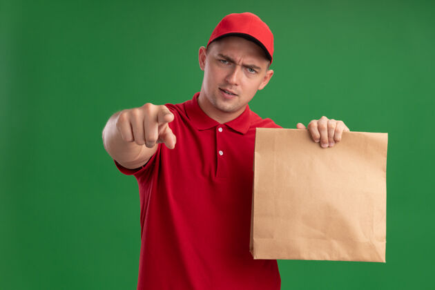 年轻人自信的年轻送货员穿着制服 戴着帽子 手里拿着一个纸食品包 在绿色的墙上向你们展示着与世隔绝的姿态穿着展示制服