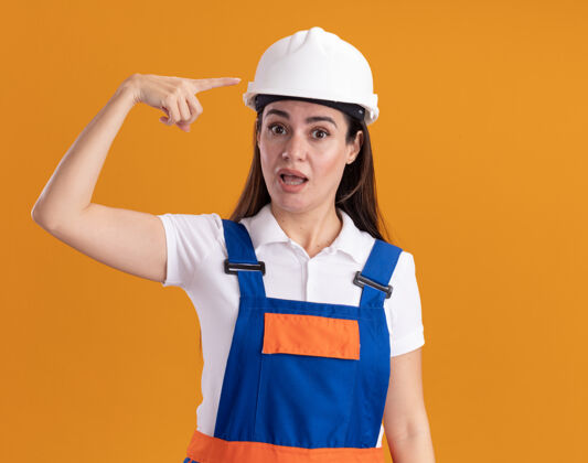 尖一个穿着制服的年轻女建筑工人惊讶地指着被隔离在橙色墙上的自己建设者制服女人
