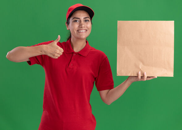 包装微笑着的年轻送货女孩穿着制服 戴着帽子 拿着纸食品包 在绿色的墙上孤立地竖起大拇指年轻人穿拇指