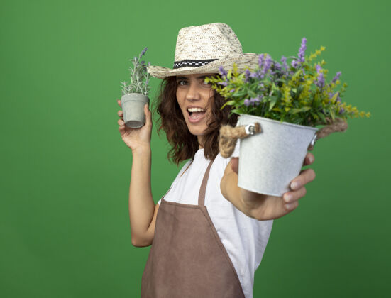 花快乐的年轻女园丁穿着制服 戴着园艺帽 在花盆里捧着花成人女人举行