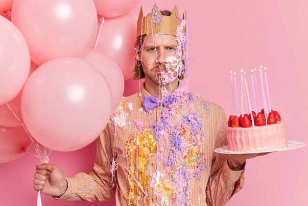 蜡烛失望沮丧沮丧的男人涂上奶油和蛇纹石喷雾准备庆祝生日的姿势与空气球和草莓蛋糕脸祝贺庆祝