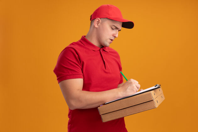 年轻人自信的年轻送货员穿着制服 戴着帽子 拿着披萨盒 在橙色墙上的剪贴板上写着什么男人制服披萨