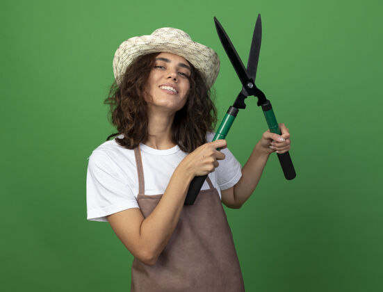 女人微笑的年轻女园丁穿着制服戴着园艺帽举起剪刀花园园艺穿