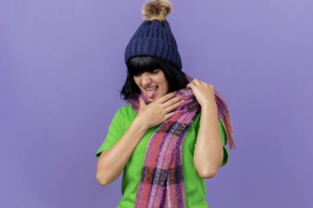 女孩恼怒的年轻生病的女人戴着冬天的帽子和围巾拿着喉咙看着隔离在紫色墙上的复制空间站脸冬天