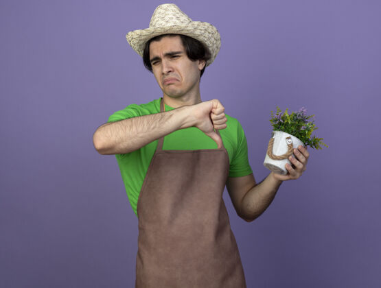 抱穿着制服的年轻男园丁戴着园艺帽 手里拿着花盆里的花 拇指朝下人花园制服