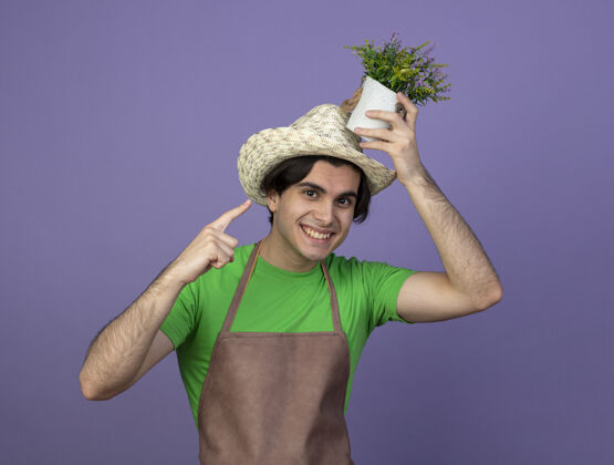 紫色身着制服的年轻男园丁微笑着 戴着园艺帽 指着花盆里的花 指着头男抱点