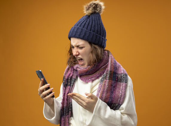 帽子愤怒的生病的年轻女孩穿着白色长袍 戴着冬天的帽子 手里拿着围巾 用手指着隔离在橙色墙上的电话电话女孩疾病