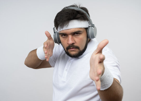 佩戴戴着头带和腕带 戴着耳机 双手孤立地站在白墙上的严格的年轻运动型男人手手持运动型