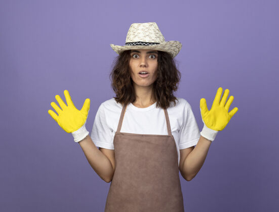 制服穿着制服的年轻女园丁惊讶地戴着园艺帽和手套摊开双手紫色人手