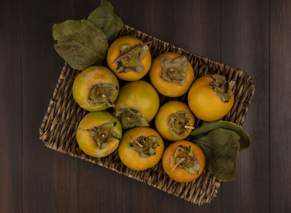 水果柳条托盘上未熟柿子果实的俯视图 木桌上有叶子盘子柿子桔子