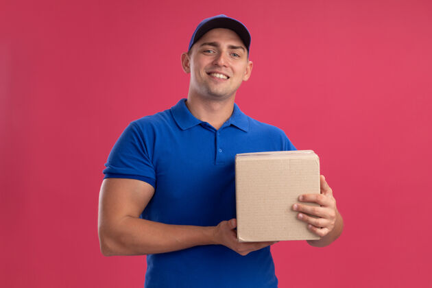 男人微笑着的年轻送货员穿着制服 粉红色的墙上有一个装帽子的盒子微笑拿着送货
