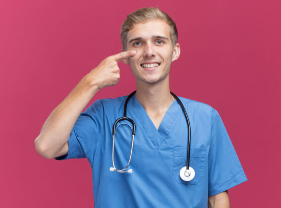 向下微笑的年轻男医生穿着医生制服 听诊器拉下眼睑隔离在粉红色的墙上戴男性听诊器