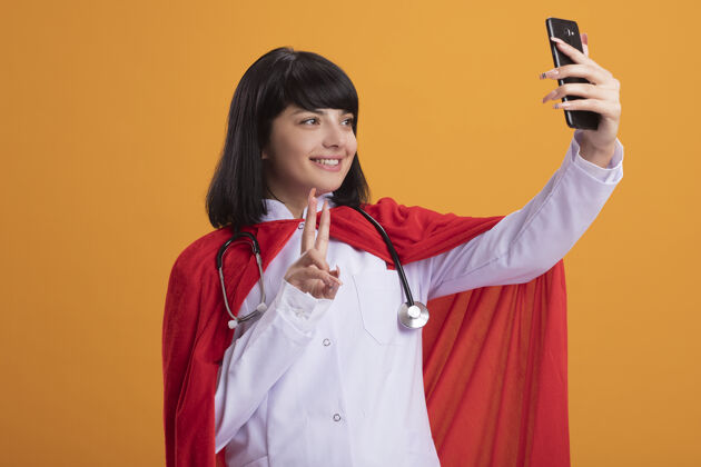 超级英雄微笑的年轻超级英雄女孩 穿着医用长袍和斗篷 戴着听诊器 自拍 展示和平姿态听诊器年轻女人