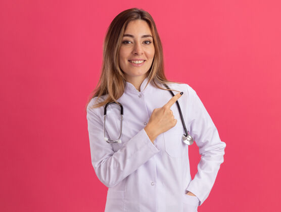 穿面带微笑的年轻女医生穿着医用长袍 听诊器指向隔离在粉红色墙上的一侧 并留有复印空间医疗微笑年轻