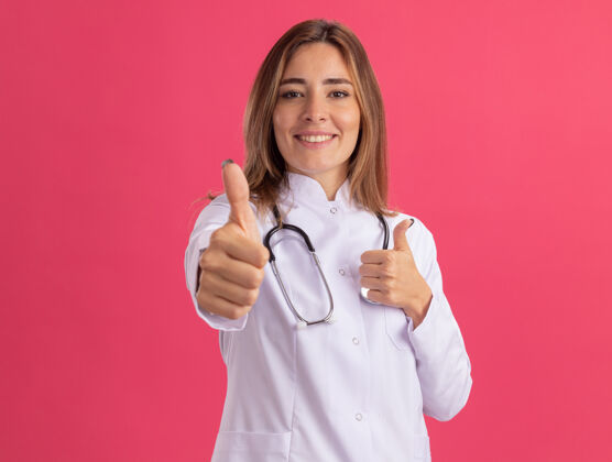 听诊器微笑着的年轻女医生穿着医用长袍 听诊器在粉红色的墙上孤立地竖起大拇指表演医疗女性