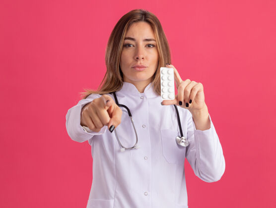 展示自信的年轻女医生 穿着医用长袍 手持听诊器的药片 在粉红色的墙上显示你的姿势穿年轻自信