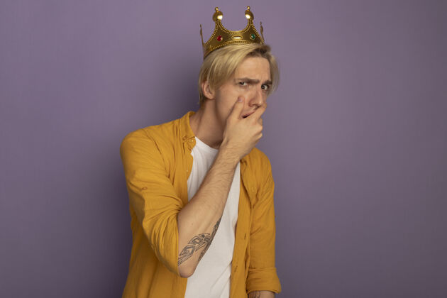 皇冠关心的年轻金发男子穿着黄色t恤和皇冠覆盖嘴与紫色孤立的手紫色年轻关心