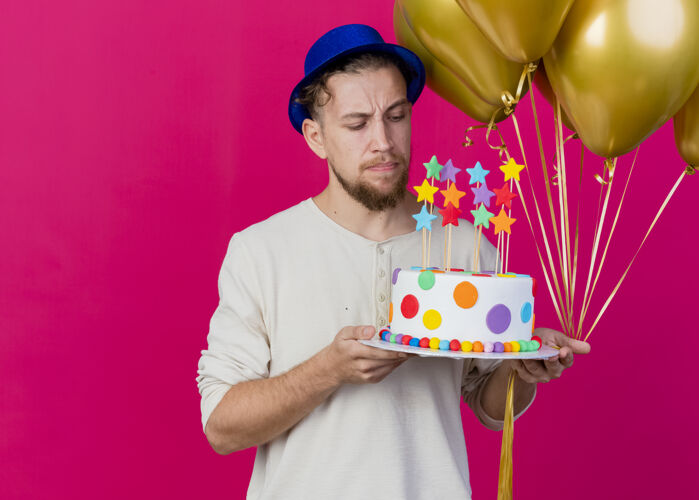 明星可疑的年轻英俊的斯拉夫党人戴着党的帽子拿着气球和生日蛋糕与明星看着蛋糕隔离在粉红色的墙壁与复制空间聚会表情黄金