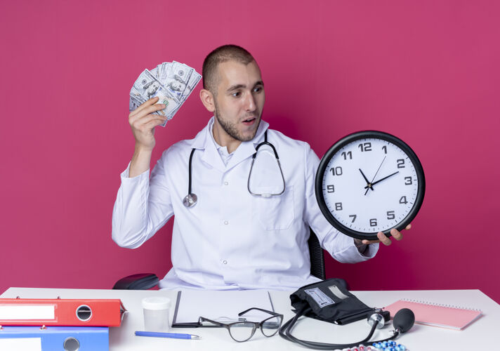 工具印象深刻的年轻男医生穿着医用长袍和听诊器坐在办公桌旁 拿着工作工具拿着时钟和钱看着粉红色的时钟长袍医生年轻