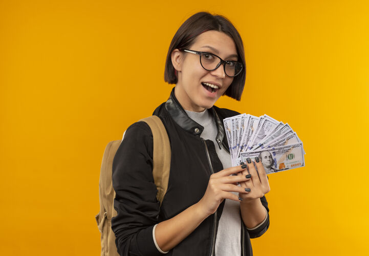 抱印象深刻的年轻女学生戴着眼镜 背着袋子拿着钱孤立在橙色背印象钱