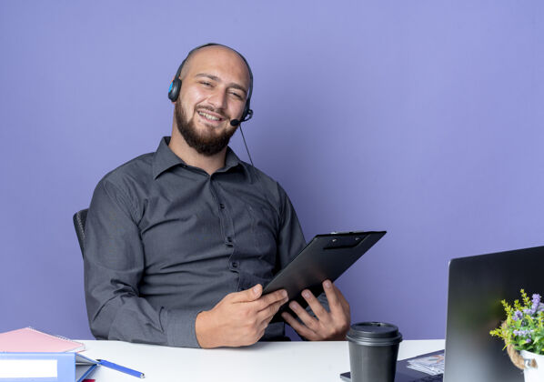 穿年轻秃顶的呼叫中心男子戴着耳机坐在办公桌旁 工作工具被隔离在紫色的背景上耳机工具电话