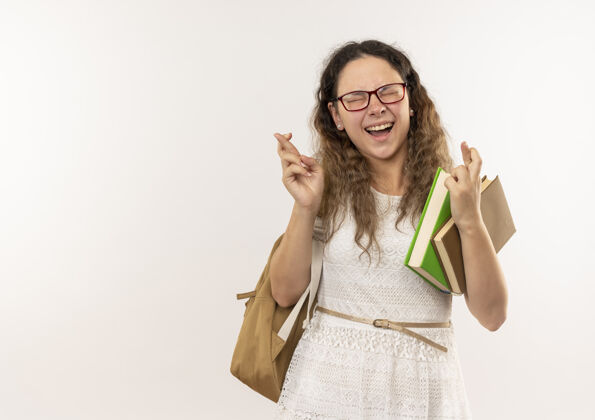 年轻快乐的年轻漂亮的女学生戴着眼镜 背着书包拿着书 闭着眼睛交叉着手指 与白色隔离眼镜闭着拿着