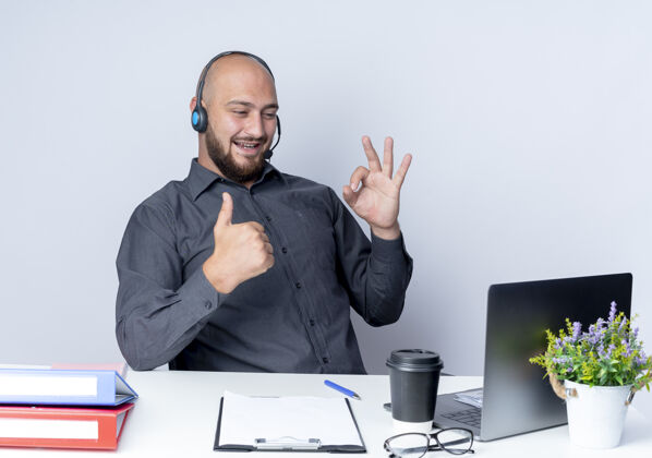 工作快乐的年轻秃头呼叫中心男子戴着耳机坐在办公桌旁 工作工具做得很好的迹象 并显示拇指在笔记本电脑上孤立的白色好耳机坐着