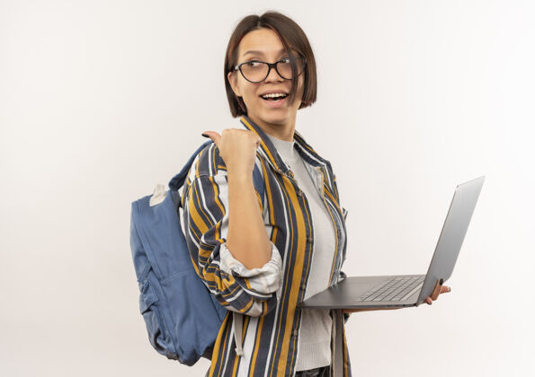 持有快乐的年轻学生女孩戴着眼镜 背着书包站在侧视图中 拿着笔记本电脑 指着后面的白色隔离带眼镜查看笔记本电脑