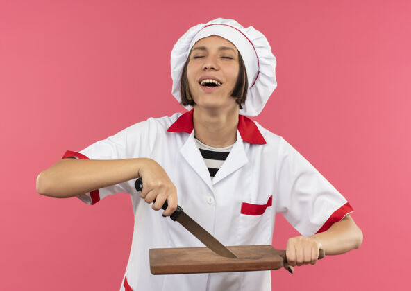 举行快乐的年轻女厨师穿着厨师制服拿着刀和砧板闭着眼睛孤立在粉红色制服粉女