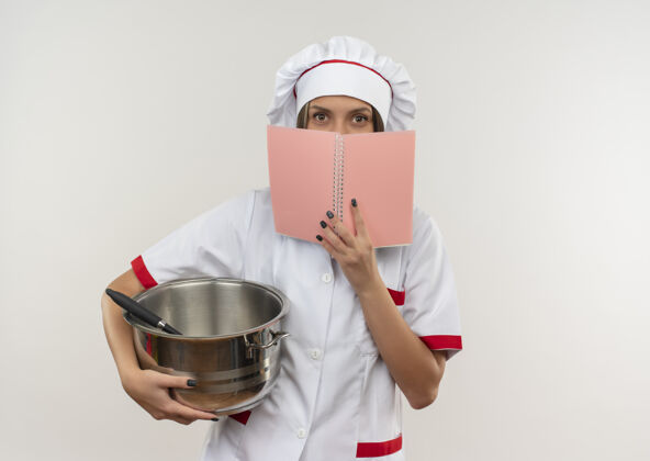 厨师穿着厨师制服的年轻女厨师拿着锅和便笺簿 从便笺簿后面看着相机 背景是白色的 有复印空间垫女相机