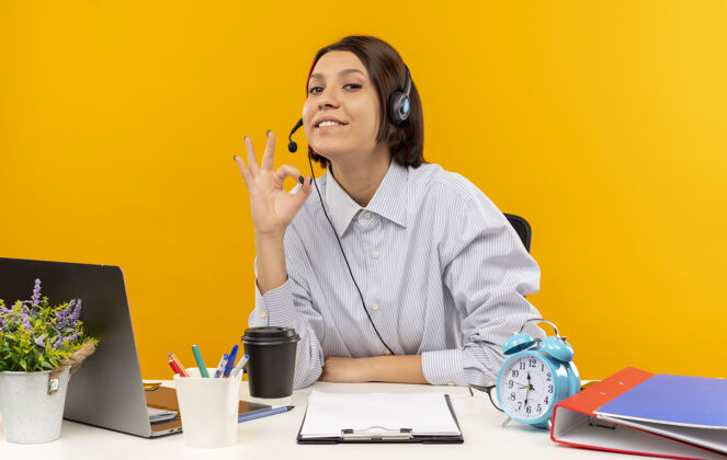 耳机高兴的年轻呼叫中心的女孩戴着耳机坐在办公桌旁做ok标志孤立的橙色背景好的年轻打电话