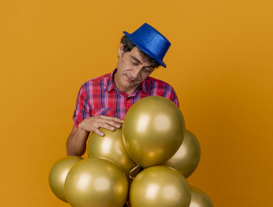 帽子一个体贴的中年人 戴着聚会帽 站在气球后面 看着气球 把手放在一个孤立的橙色墙上 留着复印空间人人人