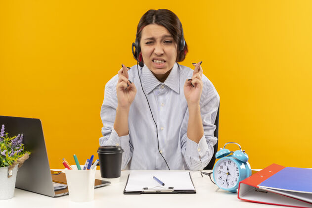 穿祝愿年轻的呼叫中心女孩戴着耳机坐在办公桌旁 工作工具交叉手指孤立在橙色背景上电话工作年轻