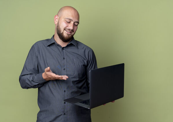 中心高兴的年轻秃头呼叫中心的男子拿着看和指着笔记本电脑孤立在橄榄绿的背景与复印空间秃头笔记本电脑请