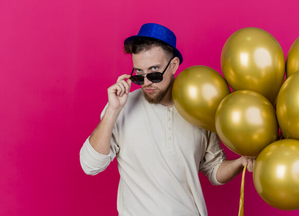 斯拉夫自信的年轻帅哥斯拉夫党家伙戴着党帽和太阳镜举行气球抓起眼镜看前面的粉红色墙上孤立的复制空间站着感觉气球