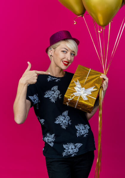 人微笑着的年轻金发派对女郎戴着派对帽拿着气球和礼盒指着礼盒看着前面孤立的粉色墙壁表情脸气球