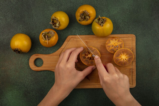 食物俯视图中的女性手切柿子水果与刀在木制厨房板上的绿色背景新鲜顶部刀